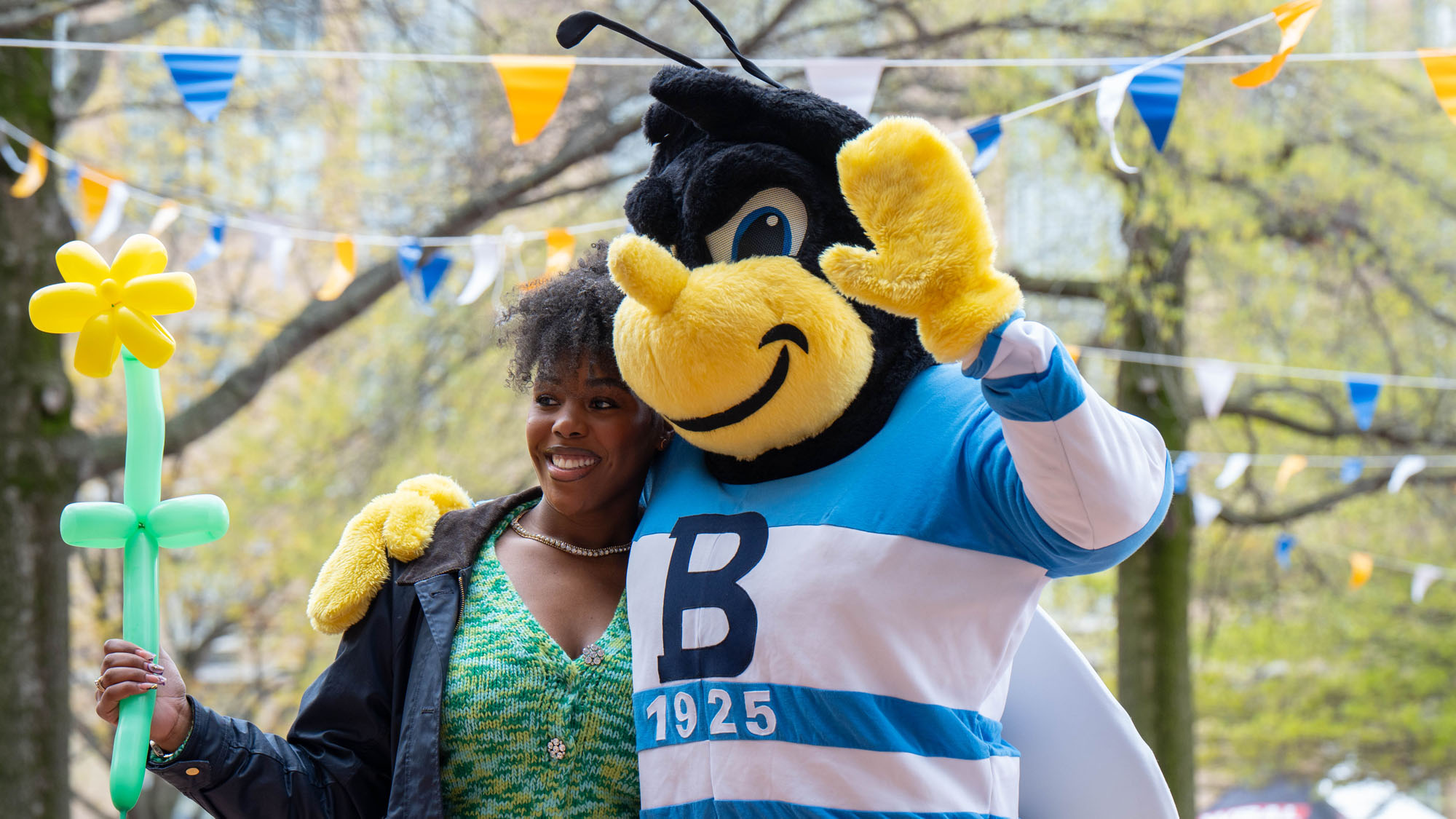 Eubie the bee mascot waves with a student holding a balloon shaped like a flower. 